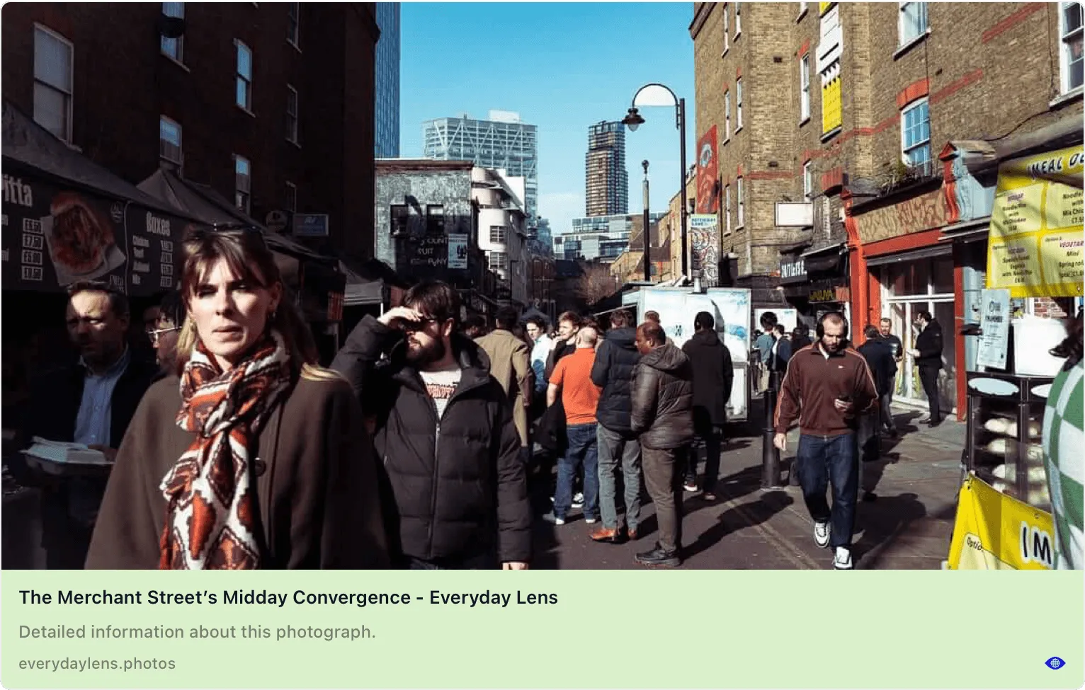 WhatsApp Open Graph preview showing a street photography from Everyday Lens. The preview displays a photograph titled The Merchant Street's Midday Convergence, featuring a busy urban street scene with pedestrians walking between brick buildings. Modern skyscrapers are visible in the background under clear blue sky. The interface shows the website title everydaylens.photos and an eye icon indicating the image can be viewed.